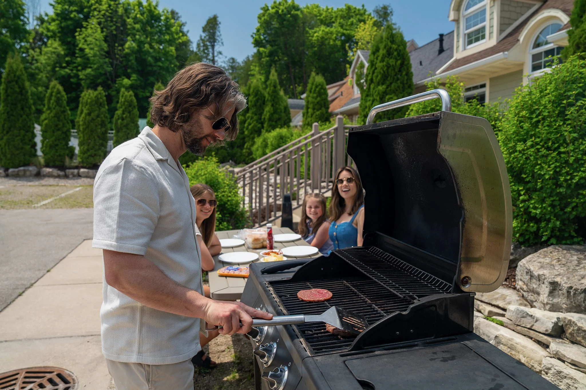 Man grilling burgers outdoors with family nearby.